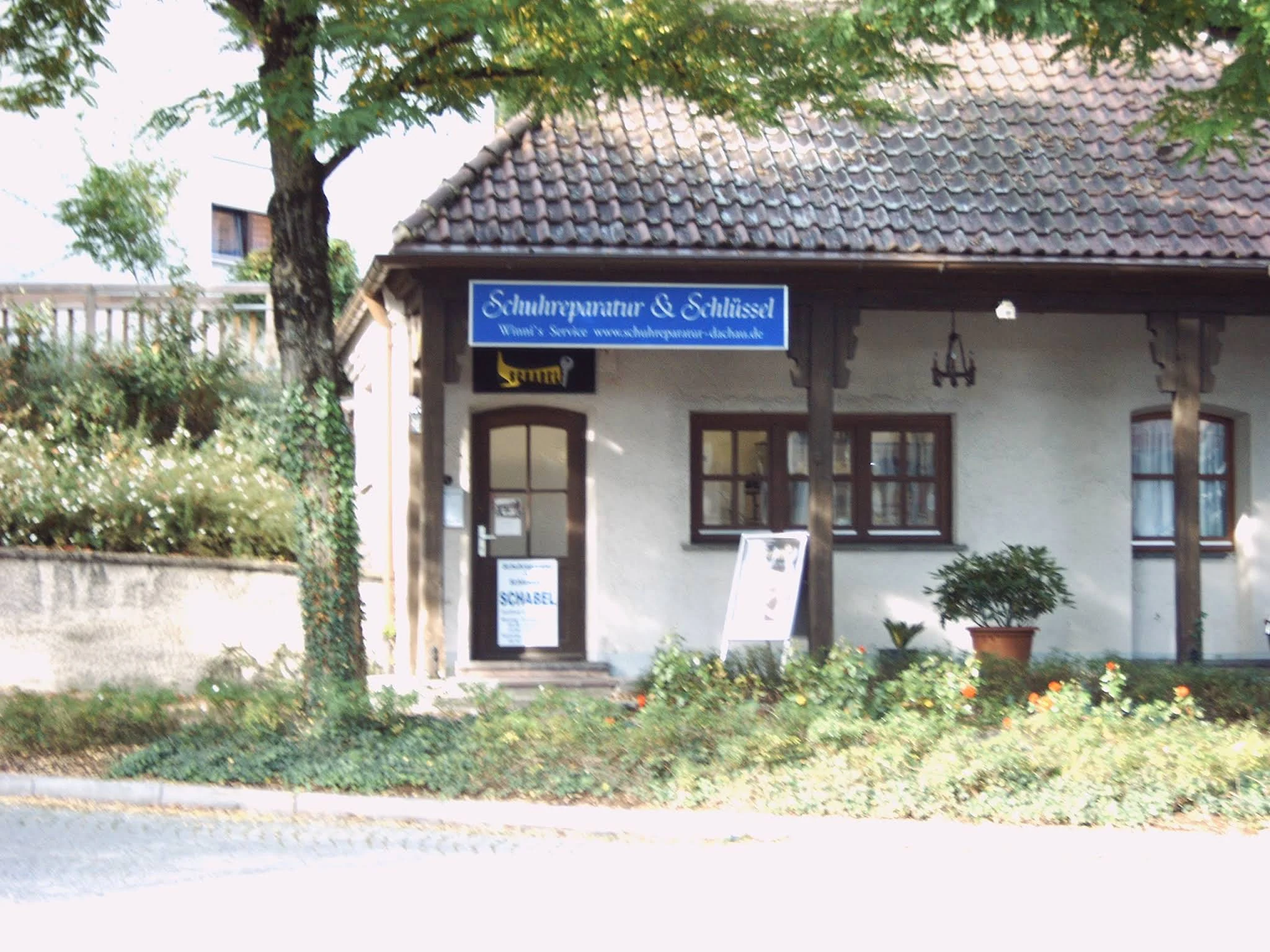 Das Geschäft Schabel in einem kleinen Haus mit Ziegeldach, Bäumen und blauem Ladenschild